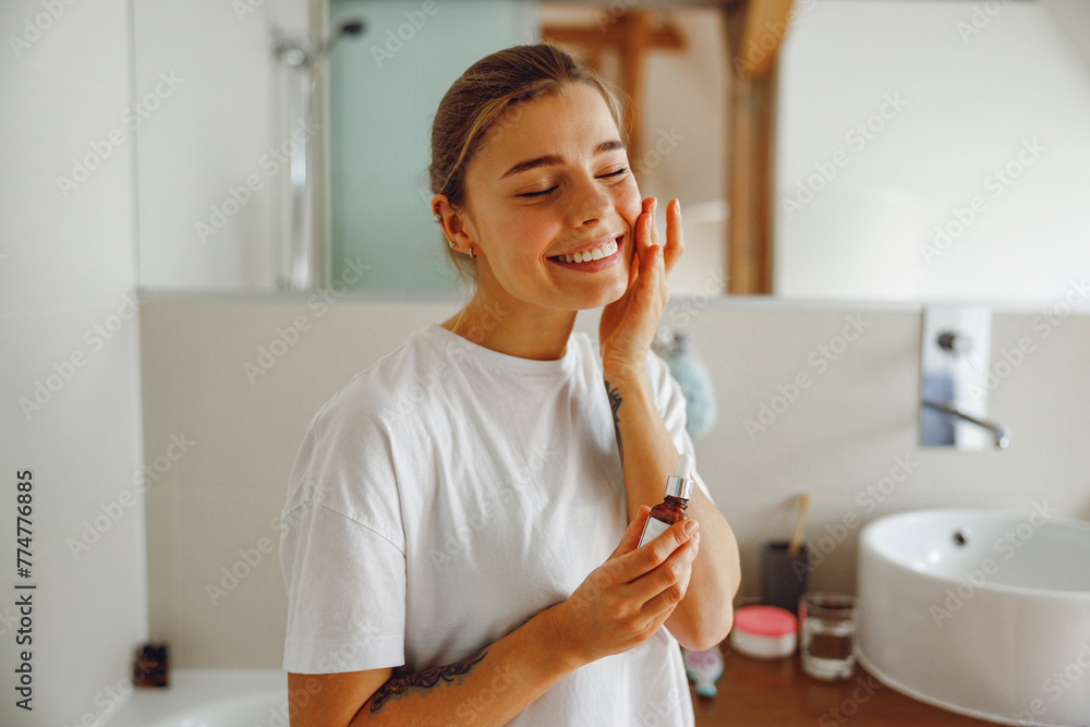 Beautiful young woman applying cosmetic serum onto her face in bathroom. Facial Skincare Beautiful young woman applying cosmetic serum onto her face in bathroom. Facial Skincare