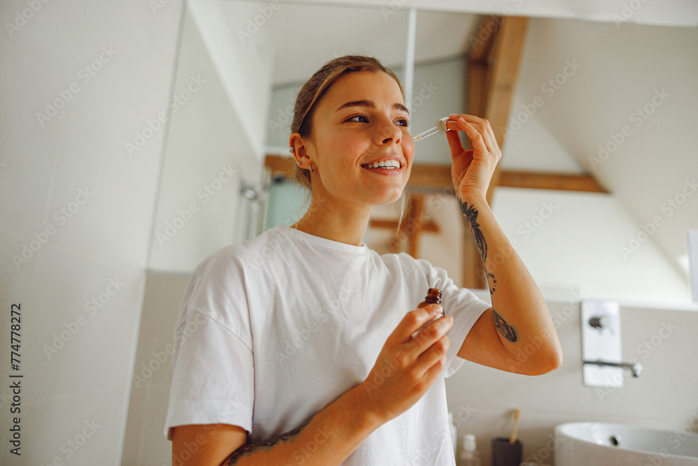Attractive female applying serum on face moisturizing and caring for skin standing in bathroom Attractive female applying serum on face moisturizing and caring for skin standing in bathroom
