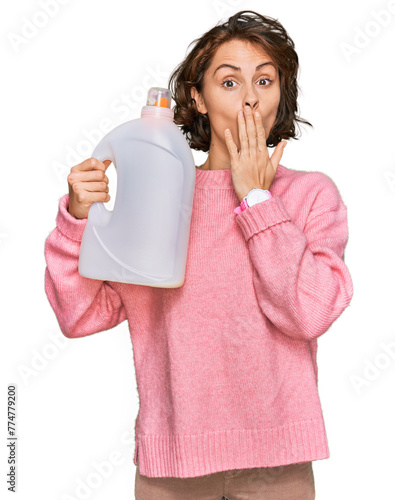 Young hispanic woman doing laundry holding detergent bottle covering mouth with hand, shocked and afraid for mistake. surprised expression