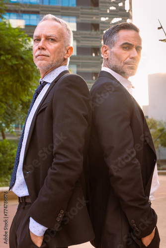 Vertical portrait two successful businessmen standing back to back looking at camera 