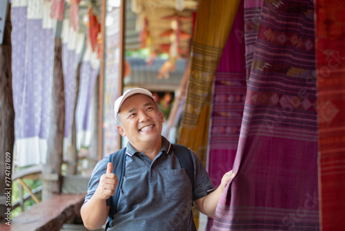 Asian middle-aged man in white cap and t-shirt, thumbinb up and holding handmade woven fabric which hanging and showing for tourists in local shop, soft focus, happiness of people concept. photo