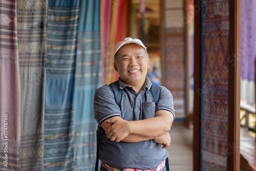 Asian middle-aged man in white cap and t-shirt standing and crossing one's arm in woven fabric shop which decorated with various fabric hanging, self confident people concept. photo