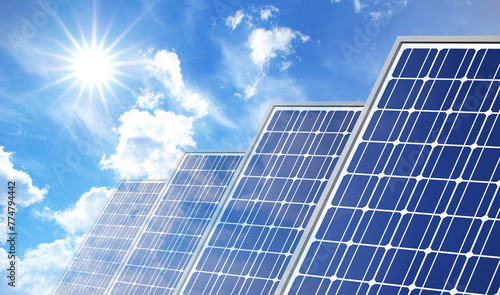 Solar Panels on Blue Sky with Sun Flares. Series of solar panels tilt upwards towards the clear blue sky, where the sun shines brightly casting a flare. Perfect day for harnessing solar energy.