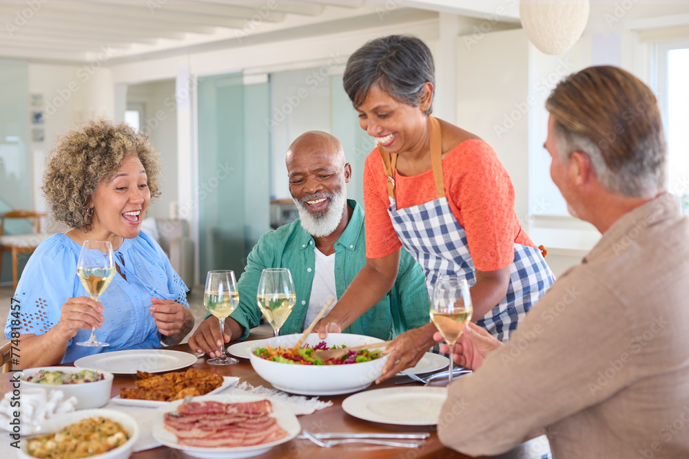 Group Of Smiling Mature Friends At Home Relaxing Meeting For Lunch And Wine Together As Woman Serves Group Of Smiling Mature Friends At Home Relaxing Meeting For Lunch And Wine Together As Woman Serves