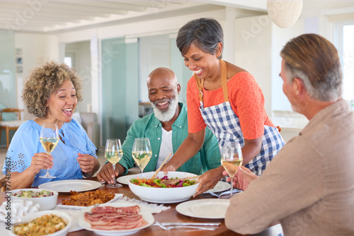 Group Of Smiling Mature Friends At Home Relaxing Meeting For Lunch And Wine Together As Woman Serves