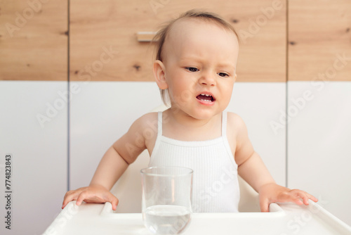 Unhappy baby sitting in a baby chair in the kitchen. Child drinks water. Feeding and childhood