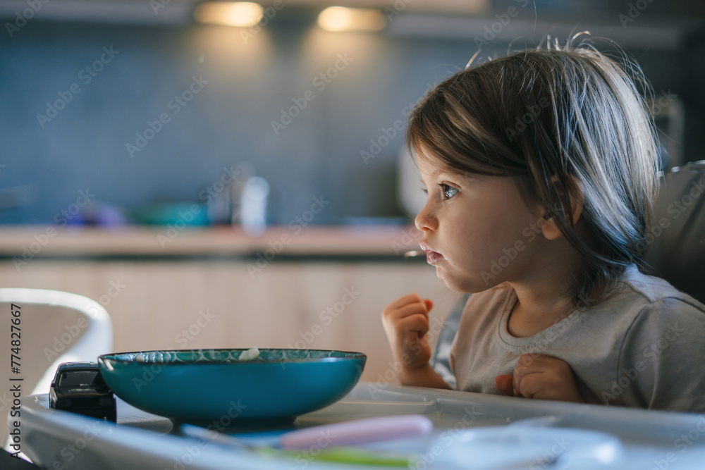 Little kid girl eating her morning porridge rice herself, sitting in baby chair in kitchen. Tasty meal, good appetite. Little kid girl eating her morning porridge rice herself, sitting in baby chair in kitchen. Tasty meal, good appetite.