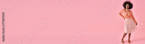 elegant african american woman in peach blouse and midi skirt posing on pastel pink backdrop, banner