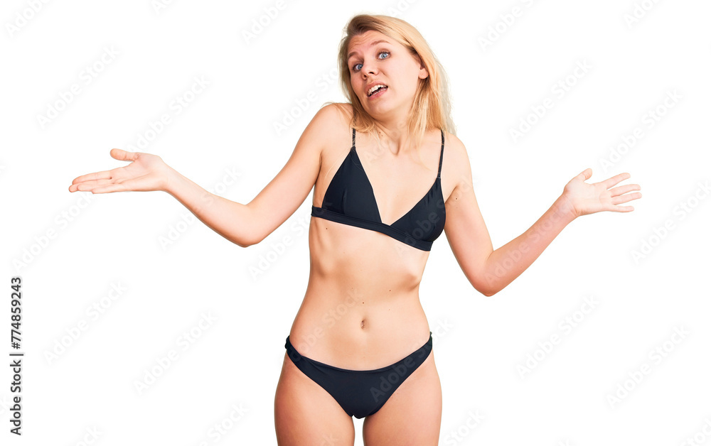 Young beautiful blonde woman wearing bikini clueless and confused expression with arms and hands raised. doubt concept. Young beautiful blonde woman wearing bikini clueless and confused expression with arms and hands raised. doubt concept.