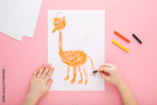 Little child girl hand holding wax crayon and drawing orange cute giraffe shape on white paper on pastel pink table background. Closeup. Point of view shot. Children creativity time. Top down view.