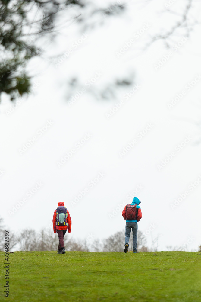 Two people are walking in a field with backpacks. The sky is cloudy and the grass is green Two people are walking in a field with backpacks. The sky is cloudy and the grass is green