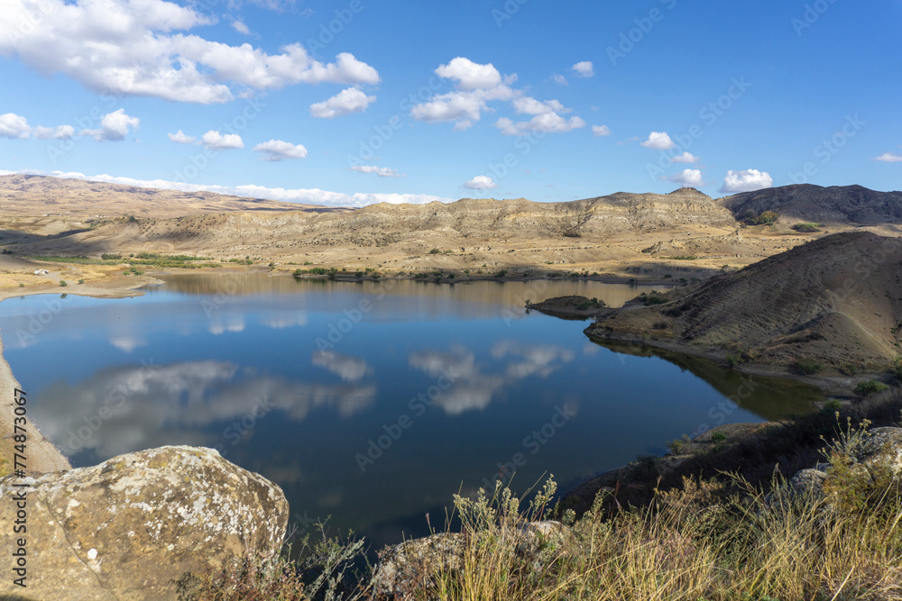 Beautiful view of the lake and mountains. Bright blue sky. Clouds are reflected in the water. Dry grass. Mravaltskaro Reservoir in Georgia.