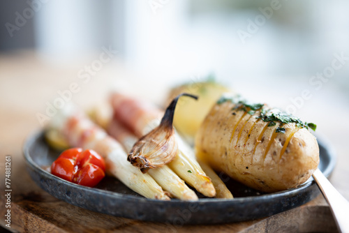 Fried white asparagus wrapped in bacon with herbed potatoes and tomatoes on black ceramic plate Close-up with short depth of field.