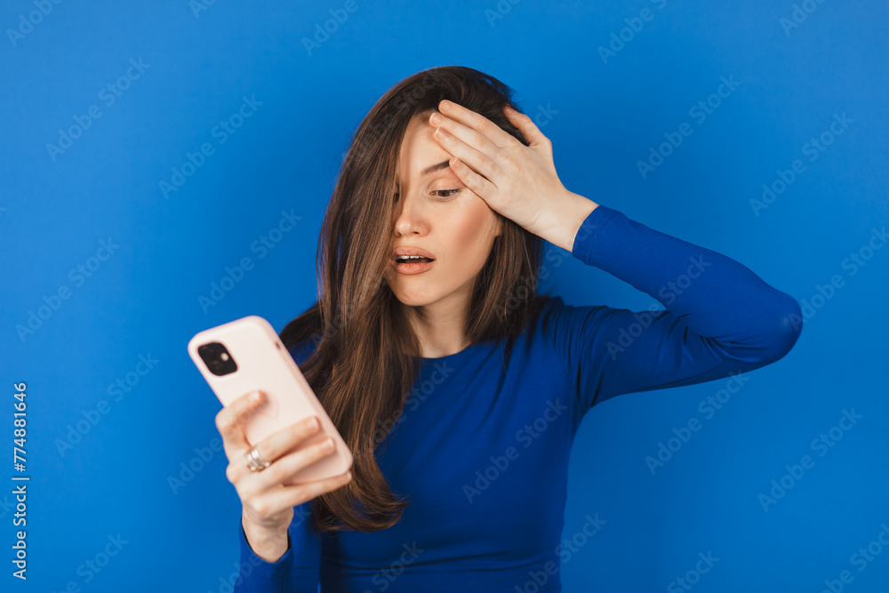 Mad and shock brunette woman make facepalm using mobile phone isolated on blue background. Yelling unhappy woman hold mobile phone. Disappointed sad upset lady horrified impressed news. WTF. Oh no. Mad and shock brunette woman make facepalm using mobile phone isolated on blue background. Yelling unhappy woman hold mobile phone. Disappointed sad upset lady horrified impressed news. WTF. Oh no.