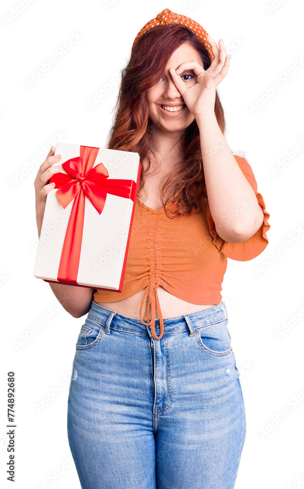 Young beautiful woman holding gift smiling happy doing ok sign with hand on eye looking through fingers Young beautiful woman holding gift smiling happy doing ok sign with hand on eye looking through fingers