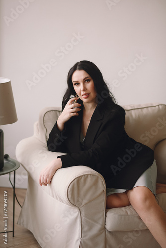 Attractive brunette woman in black jacket and blue nightdress sitting in the chair and holding perfume in her hand while posing in the studio. Space for text. Beauty and fashion