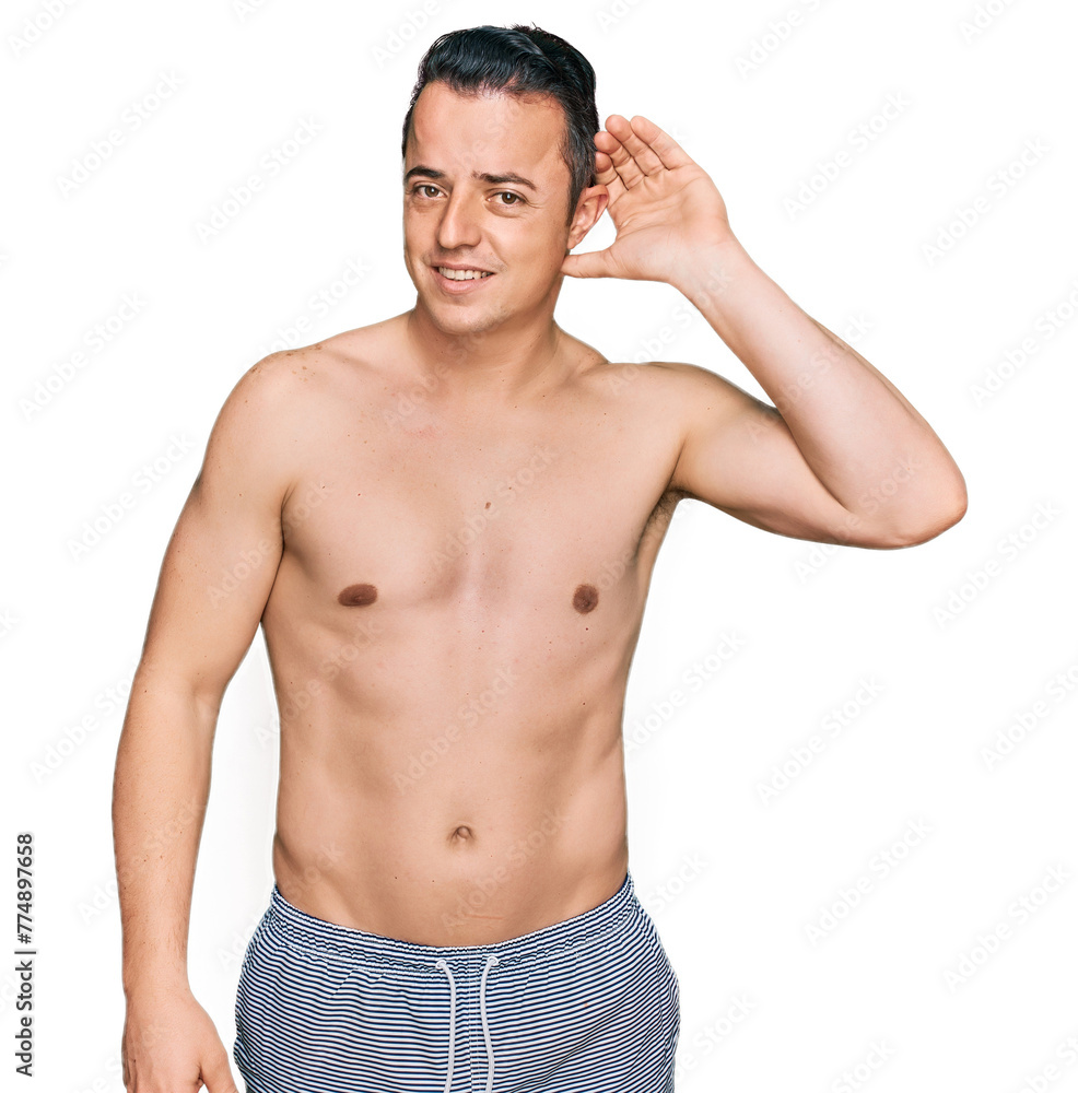 Fototapeta premium Handsome young man wearing swimwear shirtless smiling with hand over ear listening an hearing to rumor or gossip. deafness concept.