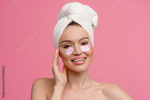 smiling h woman after shower on pink background with bath towel doing morning skincare routine using eye patches