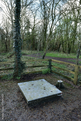 Large square metal manhole cover located in a wooded area fenced off from a footpath