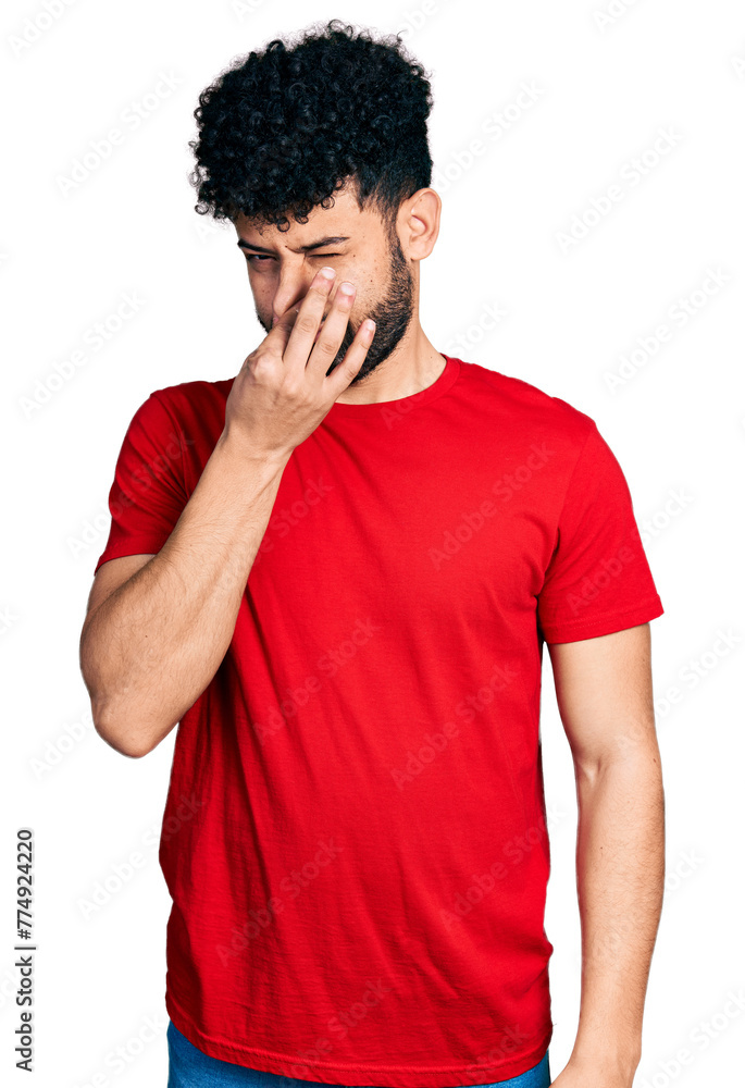 Young arab man with beard wearing casual red t shirt smelling something stinky and disgusting, intolerable smell, holding breath with fingers on nose. bad smell Young arab man with beard wearing casual red t shirt smelling something stinky and disgusting, intolerable smell, holding breath with fingers on nose. bad smell