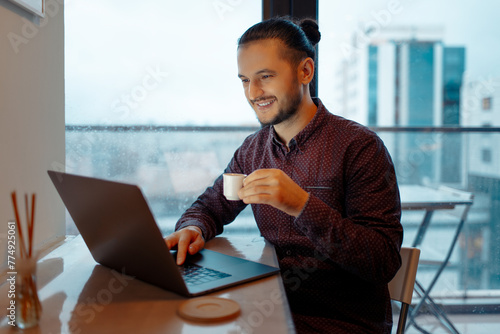 Portrait of handsome man working at laptop, typing on keyboard while holding a small cup of espresso coffee on background of panoramic windows.