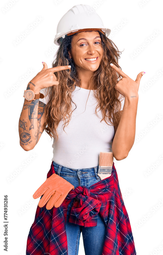 Young hispanic woman with tattoo wearing hardhat and builder clothes smiling cheerful showing and pointing with fingers teeth and mouth. dental health concept. Young hispanic woman with tattoo wearing hardhat and builder clothes smiling cheerful showing and pointing with fingers teeth and mouth. dental health concept.