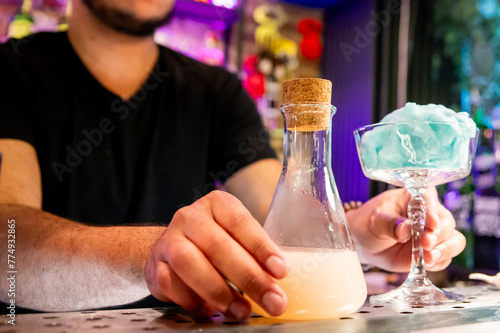 A man holds a flask with a cork stopper  next to a glass emitting blue smoke in a bar setting.