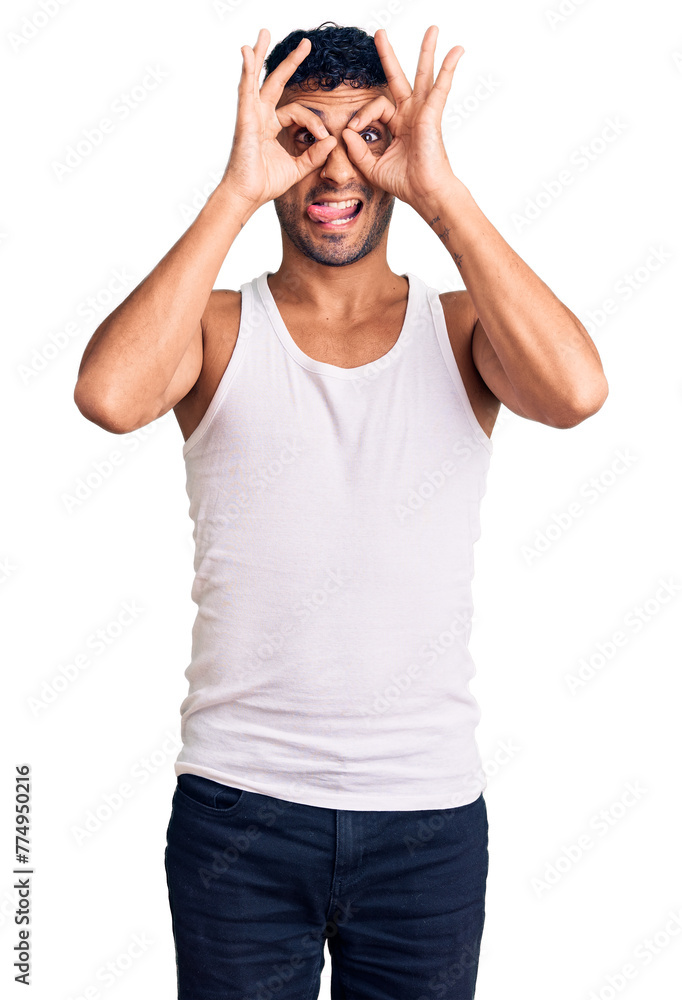 Young hispanic man wearing casual clothes doing ok gesture like binoculars sticking tongue out, eyes looking through fingers. crazy expression. Young hispanic man wearing casual clothes doing ok gesture like binoculars sticking tongue out, eyes looking through fingers. crazy expression.