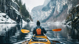 rear view of a man paddling a kayak in winter on a mountain lake surrounded by snow-covered rocks