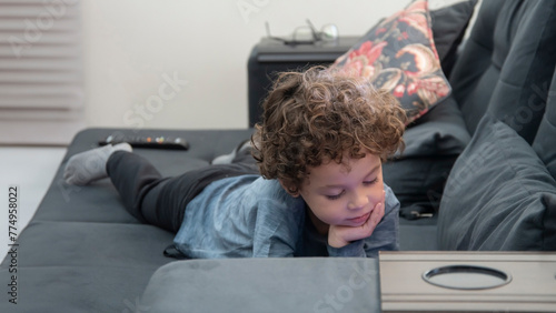 Criança com cabelos cacheados com expressão pensativa brincando de jogos no celular deitado no sofa, pensativo, ideia photo