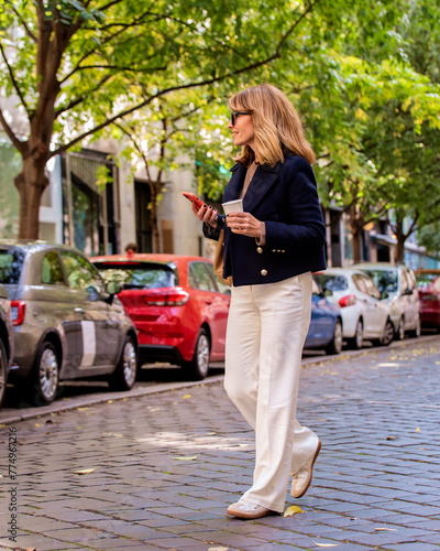 Full length of an attractive mid aged woman holding paper coffee cup and her mobile phone in her hand and walking on the city street