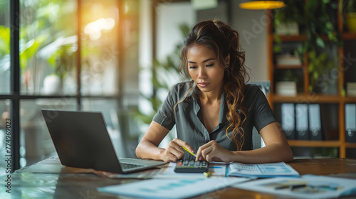 A latina woman is sitting at a desk with a laptop and a calculator, working on finances 