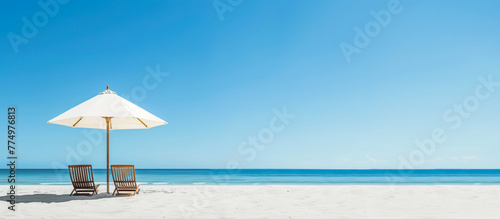 White sandy beach with two wooden lounge chairs under a large umbrella, clear blue sky and calm turquoise sea creating a serene vacation atmosphere.