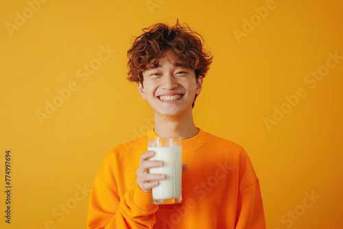 Young smiling fun happy Japanese man wear orange sweatshirt casual clothes, isolated on yellow studio background, hold in hand glass drink regular milk. 