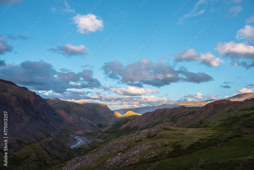 Mountain and hill tops illuminated by sunset light. Rocks silhouettes and gold sunrise colors. Golden sunlight on hills under clouds of sunset tones in blue sky. Shadows of clouds in mountain valley.
