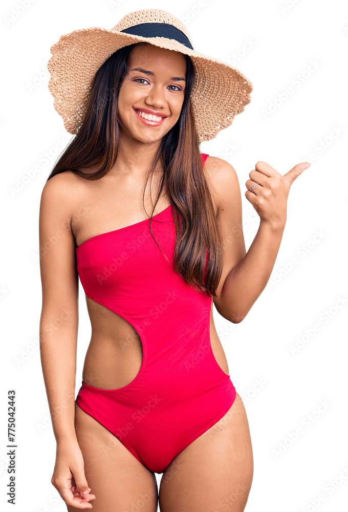 Young beautiful latin girl wearing swimwear and summer hat smiling with happy face looking and pointing to the side with thumb up. Young beautiful latin girl wearing swimwear and summer hat smiling with happy face looking and pointing to the side with thumb up.