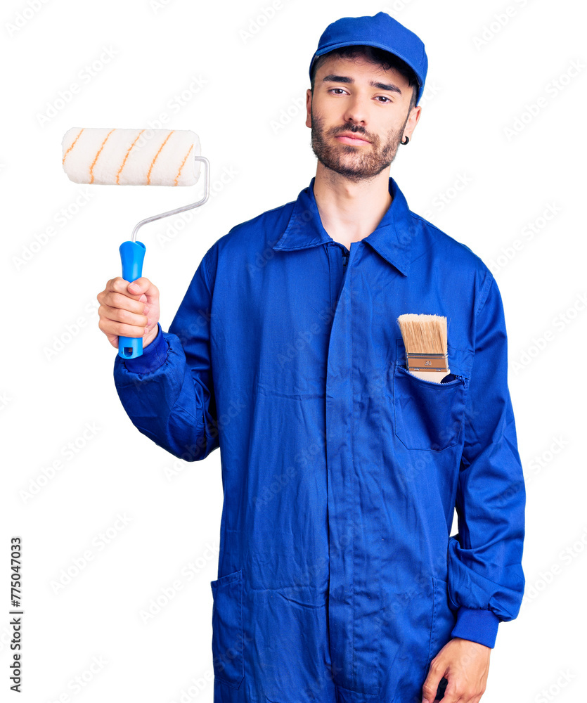 Young hispanic man wearing painter uniform holding roller thinking attitude and sober expression looking self confident