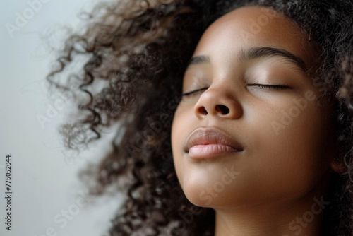 Photo of a beautiful young African American teenage girl with black short wavy hair inhaling air with her eyes closed against a light background.