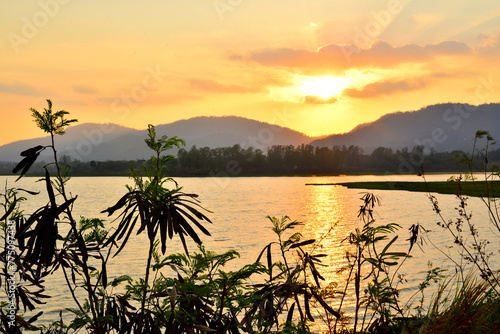 A serene sunset over a calm lake, surrounded by silhouetted mountains and foregrounded by darkened vegetation, evoking a sense of peaceful solitude.