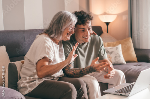 Video call concept. Happy senior retired woman sitting on sofa with young nephew waving hands video calling by laptop using technology for online webcam connection with distant family or friends photo