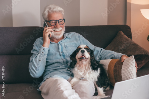 Senior man talking on mobile phone sitting on sofa with his cavalier king charles dog. Elderly male communicate with smart phone with his best friends nearby