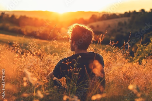 Silhouetted male figure seated in a lush field, basking in the warm, golden light of a setting sun, evoking a sense of peace and contemplation photo