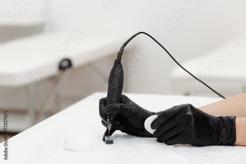 A professional tattoo artist, wearing black gloves, prepares for a session with a tattoo machine on a clean, white surface.