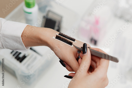 An eyelash extension specialist prepares for the procedure. glued two strips of artificial eyebrows to one hand and holds tweezers in the other. Background is light and blurry background close-up