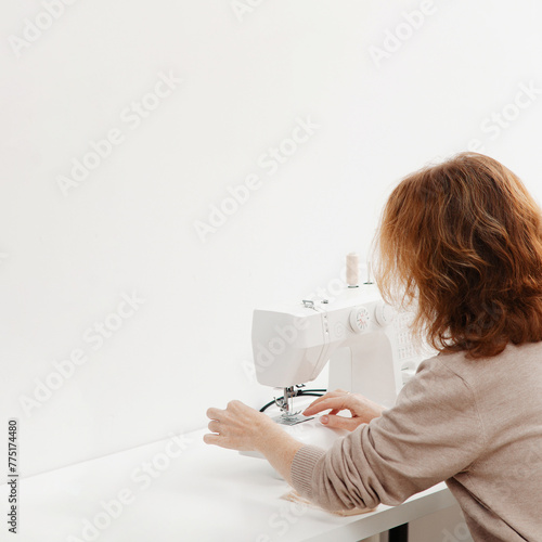 A woman with red hair, dressed in a light-colored sweater, is seen from behind diligently working at a sewing machine.