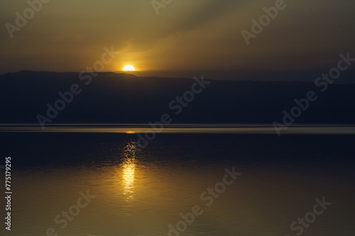 Jordan. Dead Sea. Calm at sea. Sunset on Dead Sea coast. Orange sun is reflected in water. There is light path in water from reflection of sun. Opposite bank with mountains is Israeli territory.