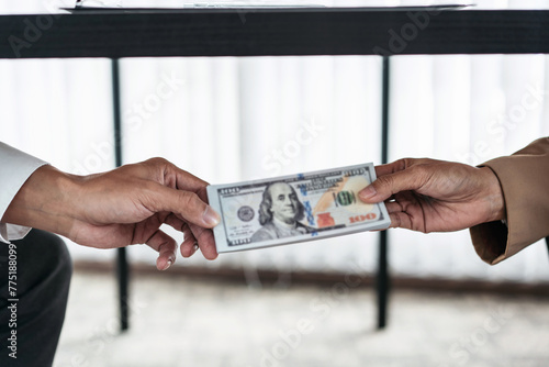 A person is giving a stack of dollar bills to another person under table. The person receiving the money is wearing a suit