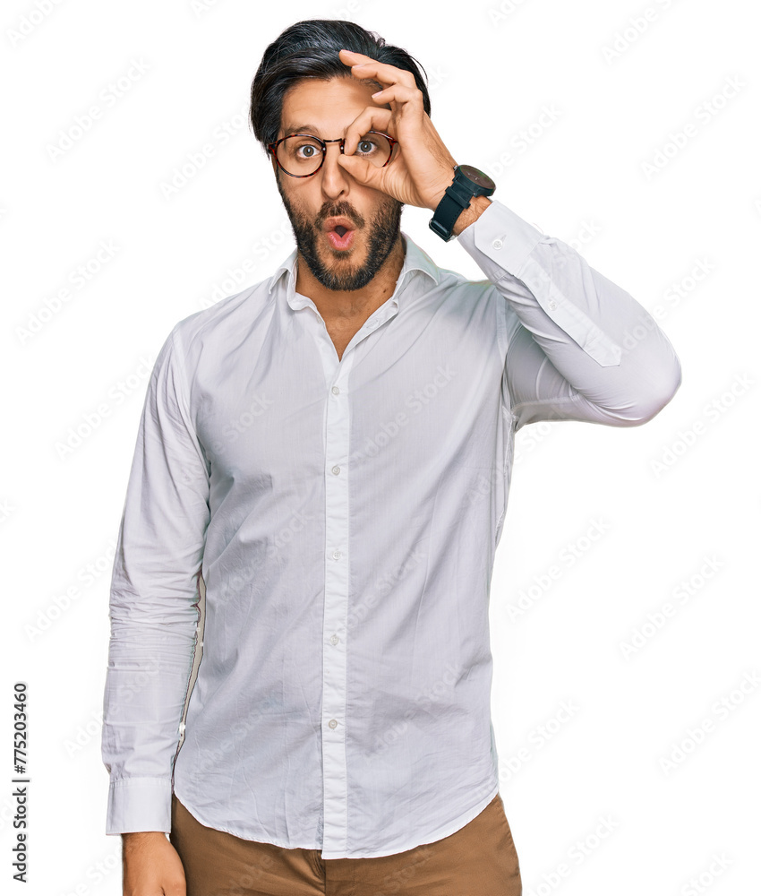 Naklejka premium Young hispanic man wearing business shirt and glasses doing ok gesture shocked with surprised face, eye looking through fingers. unbelieving expression.