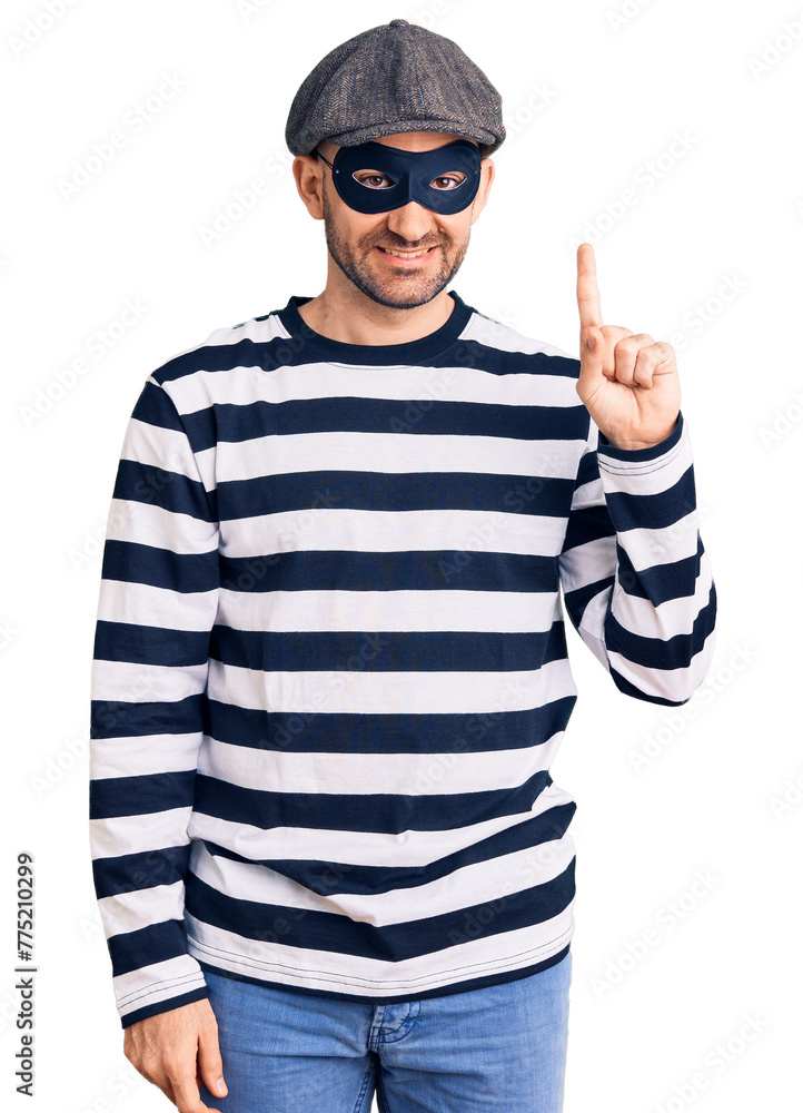 Young handsome man wearing burglar mask showing and pointing up with finger number one while smiling confident and happy. Young handsome man wearing burglar mask showing and pointing up with finger number one while smiling confident and happy.