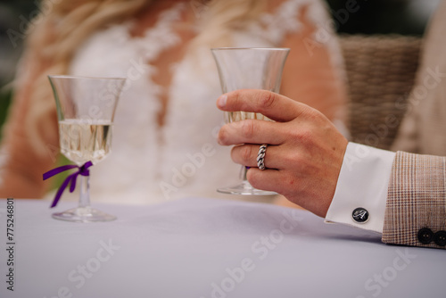 Valmiera, Latvia - Augist 13, 2023 - Close-up of a bride and groom's hands holding champagne glasses at a wedding, showcasing rings, elegant attire, and a hint of a floral bouquet.